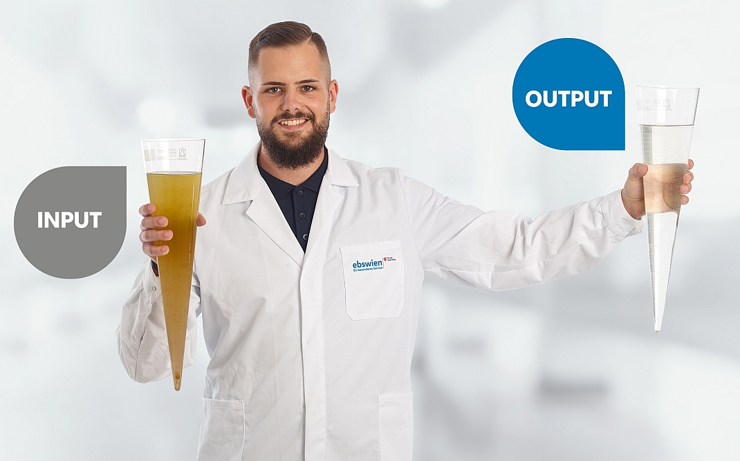 Das Foto zeigt einen Mitarbeiter der Wiener Kläranlage im weißen Labormantel, der zwei Imhof-Trichter hält, die den braun gefärbten Zulauf (beschriftet mit "Input") beziehungsweise den farblosen Ablauf (beschriftet mit "Output") enthalten. Das Bild zeigt den 20 Stunden dauernden Abwasserreinigungsprozess in der Wiener Kläranlage somit auf einen Blick. © Christian Houdek
